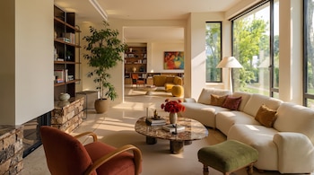 Salón con sofá, sillón, puf, mesa de centro con flores, estantería con libros, chimenea, planta, lámpara de pie y ventanales.