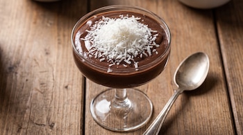 Postre de chocolate en copa de vidrio, cubierto con coco rallado, sobre mesa de madera, junto a una cuchara plateada.