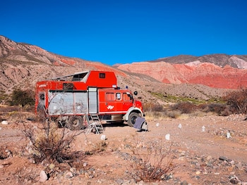 Motorhome rojo transformado de camión de bomberos en paisaje árido. Montañas rojizas, arbustos secos, suelo rocoso y cielo azul. Escalera y tela junto al vehículo