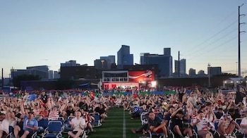 Gran multitud de personas levanta los brazos y celebra en un campo verde, con edificios urbanos y una pantalla de "Coca-Cola Fan Zone" al fondo al atardecer