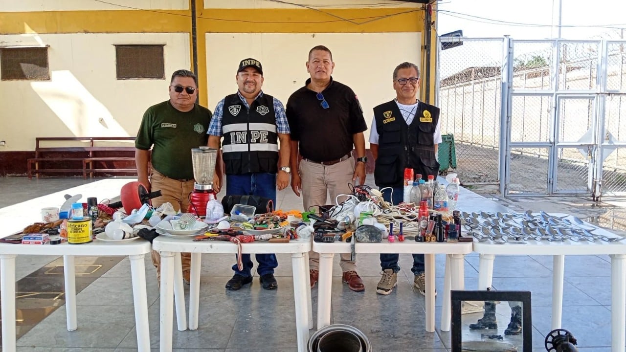 Cuatro agentes del Instituto Nacional Penitenciario (INPE) posan junto a mesas con artículos prohibidos incautados durante un operativo de seguridad en el Establecimiento Penitenciario Chiclayo para fortalecer el control y la disciplina. (Foto: INPE)