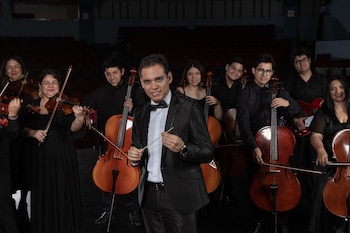 Un director con esmoquin y batuta posa al frente de varios músicos que sostienen violines, violonchelos y una guitarra eléctrica