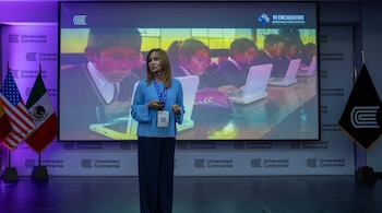 Una mujer en el centro de un escenario hablando con micrófono de diadema, frente a una pantalla que muestra niños usando laptops. A un lado, banderas