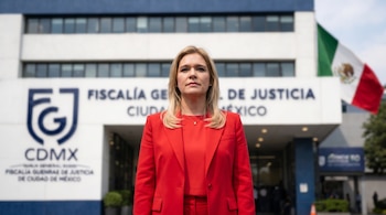 Una mujer rubia con un traje rojo mira al frente, de pie ante el edificio de la Fiscalía General de Justicia de la CDMX y una bandera mexicana ondeando.