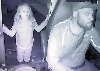 Fotografía de baja luz de una niña rubia de pie dentro de un vehículo, al lado de un hombre barbudo con gorra y sentado en el asiento del conductor