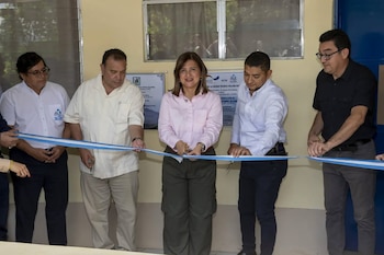 Cinco personas, una mujer en el centro corta una cinta azul y blanca con tijeras. Dos hombres sostienen la cinta. Fondo con pared y dos carteles informativos