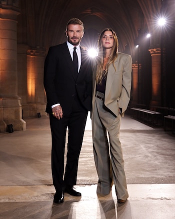 David Beckham en traje oscuro y Victoria Beckham en traje beige holgado posan de pie en una sala con arcos de piedra y luces de fondo