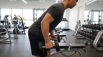 Hombre de complexión atlética con camiseta gris y pantalón corto negro sostiene dos mancuernas en un gimnasio con equipo de pesas y espejos.