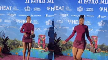 Dos mujeres con vestidos burdeos posan en una alfombra magenta. Un fondo azul con logos de Heat Latin Music Awards, Jalisco y paisaje ilustrado.