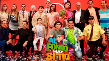 Un grupo de veinte personas posa en un escenario rojo con confeti, algunos de pie y otros arrodillados, delante del logo 'Al fondo hay sitio'
