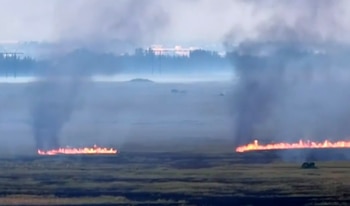 Dos líneas de fuego arden en un campo con vegetación seca. Humo gris se eleva de las llamas. El fondo muestra una línea de árboles borrosa por la bruma