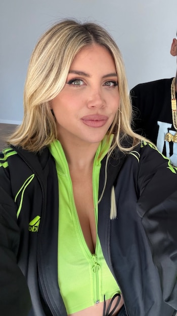 Wanda Nara vestida con chaqueta deportiva verde y negra en primer plano, con un hombre con cadenas de oro detrás