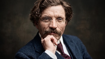 Hombre de cabello castaño con anteojos redondos, bigote, traje azul y corbata burdeos con la mano sobre su mentón.