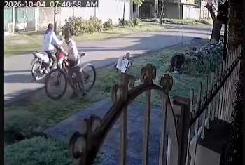 Dos personas sobre una motocicleta al lado de una persona que cae de su bicicleta en una calle pavimentada frente a una reja