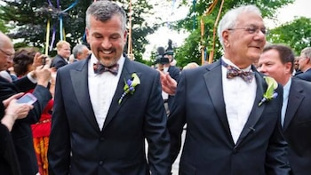 Primer plano de dos hombres adultos, uno con barba, ambos vistiendo esmoquin azul marino con pajaritas y boutonnières, sonriendo en un evento al aire libre