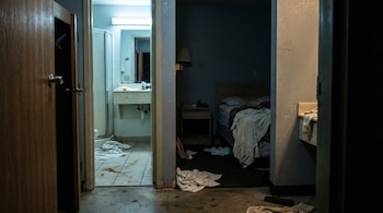Habitación de auto hotel en penumbra con baño iluminado al fondo. Se ven toallas tiradas, una cama desordenada y manchas rojizas en el suelo del baño.