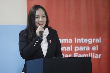 Mujer de cabello oscuro con blazer negro, blusa blanca y pin de escarapela peruana, habla en un micrófono sobre un podio. Paredes roja con texto y celeste al fondo