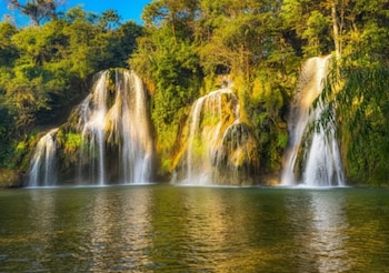 Varias cascadas de agua descienden sobre formaciones rocosas cubiertas de árboles y vegetación frondosa. En la base, un cuerpo de agua sereno