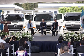 Varios funcionarios caminan junto a una hilera de camiones blancos con emblemas oficiales de Guatemala y Taiwán en un estacionamiento