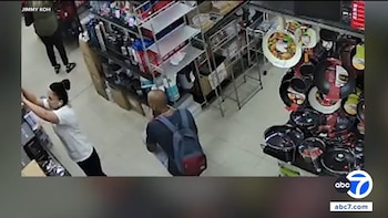 Imagen de seguridad en un comercio con estantes de utensilios de cocina, donde una mujer revisa un anaquel y un hombre con mochila permanece detrás