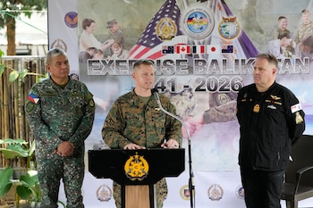 Tres oficiales militares uniformados de diferentes países, uno en el centro hablando en un podio con micrófono, flanqueado por otros dos. Detrás, un cartel del Ejercicio Balikatan con banderas