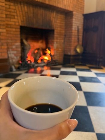 Mano sostiene taza blanca con líquido oscuro. Fondo desenfocado muestra chimenea de ladrillo con fuego, suelo a cuadros y mueble de madera