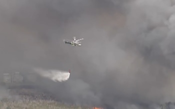 Un helicóptero cisterna vuela a baja altura, soltando una gran cantidad de agua sobre un área ardiendo con llamas y emitiendo una espesa columna de humo gris oscuro