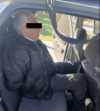 Un hombre con gafas y cabello gris, vestido con chaqueta oscura y suéter gris, sentado en el asiento trasero de un auto con la puerta abierta y las manos atadas