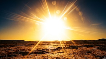 Un sol brillante emite destellos de luz sobre un terreno desértico con grietas. El cielo es azul oscuro en la parte superior y anaranjado en el horizonte