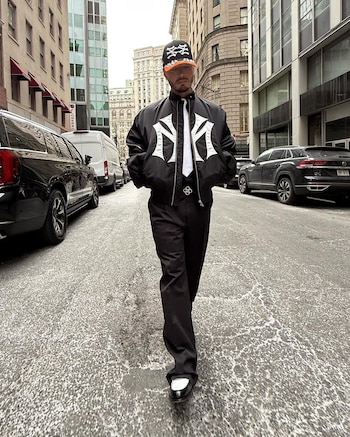 Chaqueta negra con logos blancos, pantalón negro, camisa blanca, corbata negra, zapatos bicolor y gorra negra y naranja en un look urbano y gráfico (@jbalvin)