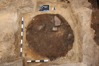 Fosa circular excavada en la tierra con piedras en el interior, dos barras de escala métrica, una flecha y un cartel de identificación