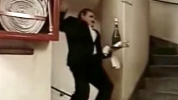 Tandarica, un hombre con esmoquin, sube unas escaleras con una pose dramática, sosteniendo una bandeja con una botella de champán y un paño blanco