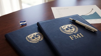 Dos carpetas azules con el logotipo del FMI, una estilográfica, un pin de El Salvador y un gráfico sobre una mesa de madera.