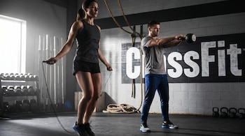 Dos personas entrenando en un gimnasio CrossFit. Una mujer salta la cuerda a la izquierda y un hombre hace kettlebell swings a la derecha. Fondo con equipo y logotipo CrossFit.