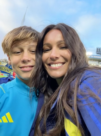 Primer plano de Pampita y su hijo Beltrán sonriendo a la cámara en un estadio de fútbol. Ella lleva una chaqueta azul y amarilla, él una turquesa
