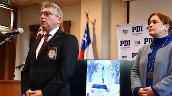 Un hombre con traje y corbata habla por un micrófono junto a una mujer. Al fondo, se ve la bandera de Chile y carteles de la PDI