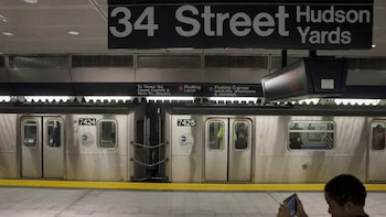 Un tren de metro plateado está estacionado en una plataforma de la estación 34 St-Hudson Yards, con un letrero grande de la estación y un cartel de ruta