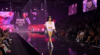 Una modelo viste un conjunto rosado y camina sobre una pasarela oscura flanqueada por espectadores y pantallas que indican PINK RUNWAY 2024.
