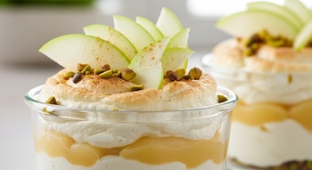 Postre en vaso con capas de crema, puré de manzana, merengue tostado, rodajas de manzana verde, pistachos y canela espolvoreada. Fondo borroso.