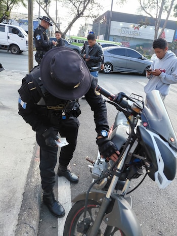 Oficial de policía de Guatemala con sombrero oscuro inspeccionando una motocicleta gris en la calle, con otros agentes y personas al fondo