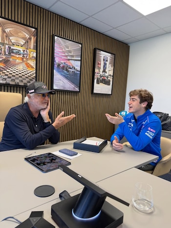 Flavio Briatore y Franco Colapinto sentados en una mesa de oficina, gesticulando durante una conversación. La pared tiene paneles de madera y pósteres de F1