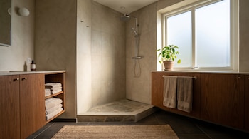 Baño moderno con ducha a nivel del suelo sin mamparas, paredes de microcemento, azulejos claros, gabinetes de madera, toallas y una planta verde en la ventana.