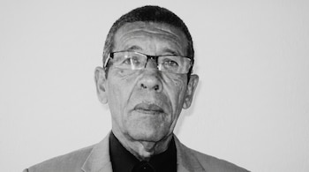 Retrato en primer plano y en blanco y negro de un hombre mayor con gafas, camisa oscura y chaqueta de traje frente a un fondo claro.