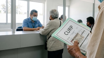 Mano de una persona sostiene una carpeta con documentos CURP y credencial INE sobre un mostrador. Un funcionario con mascarilla atiende a una persona adulta mayor.