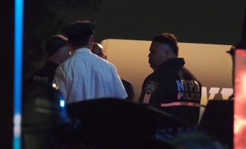 Un grupo de agentes de policía, uno con chaqueta 'NYPD Police', de noche en un lugar con luces azules y rojas y una estructura clara de fondo