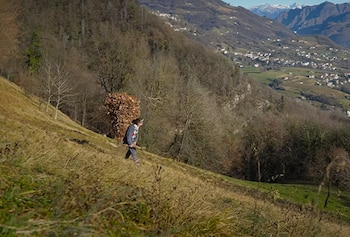 Una persona camina en una pendiente con una gran carga de madera a la espalda en un paisaje montañoso con casas al fondo