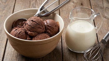 Vista cenital de un cuenco con varias bolas de helado de chocolate y una jarra de cristal llena de nata, con una cuchara para helado y un batidor.