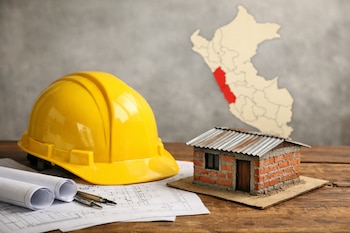 Casco amarillo sobre planos, maquetas de vivienda informal de ladrillo y techo de calamina, y mapa de Perú con Ica en rojo sobre mesa de madera.