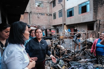 Grupo de mujeres conversando en una calle con escombros, metales retorcidos, objetos calcinados y frutas. Edificios de ladrillo al fondo