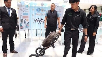 Cuatro jóvenes en una exposición; uno manipula un sistema de transporte plegable para bebés frente a un cartel "Tecnobike" con diagramas y globos de fondo